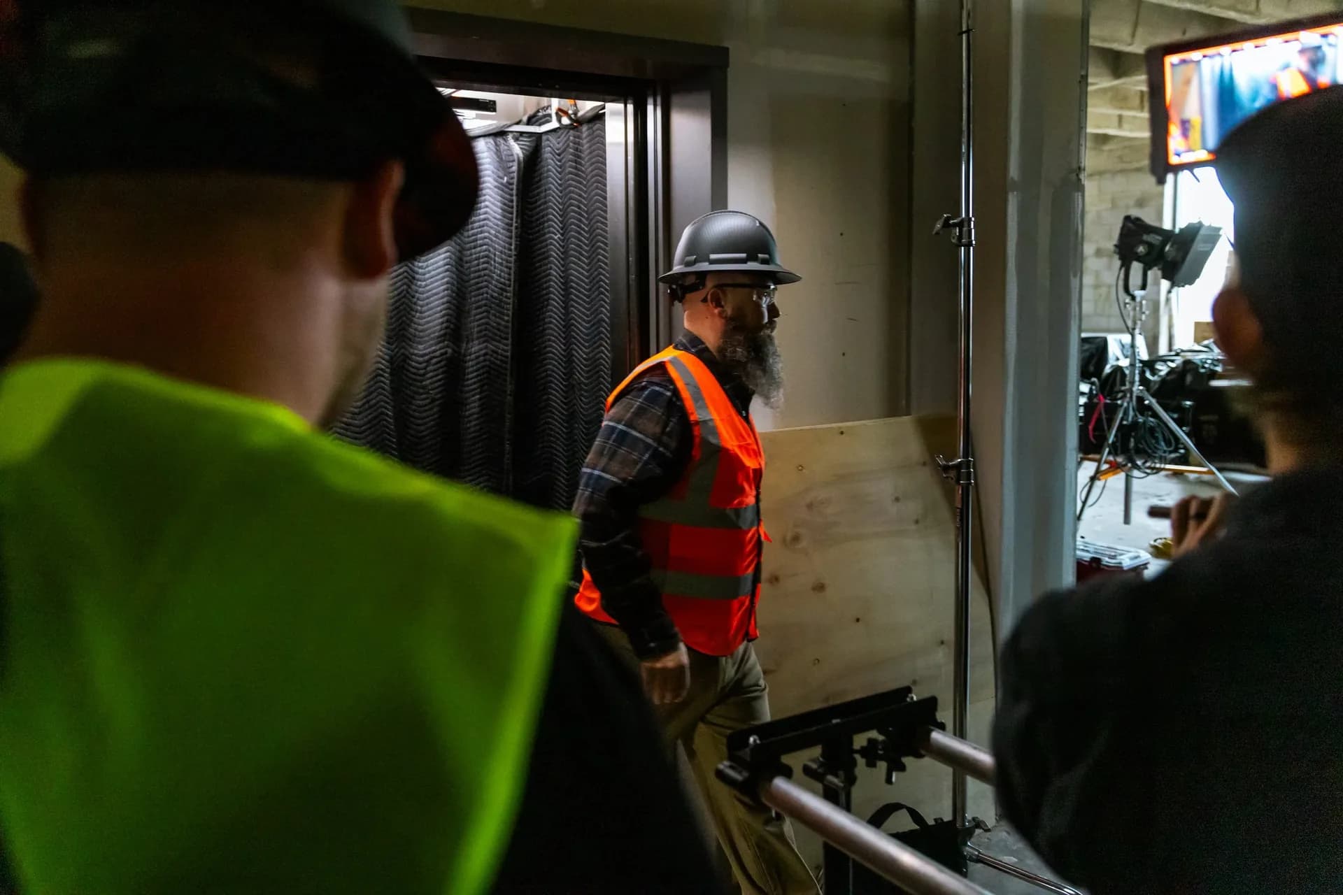 Behind the scenes at the elevator setup: a worker in a hard hat and orange vest steps out of the car past the crew