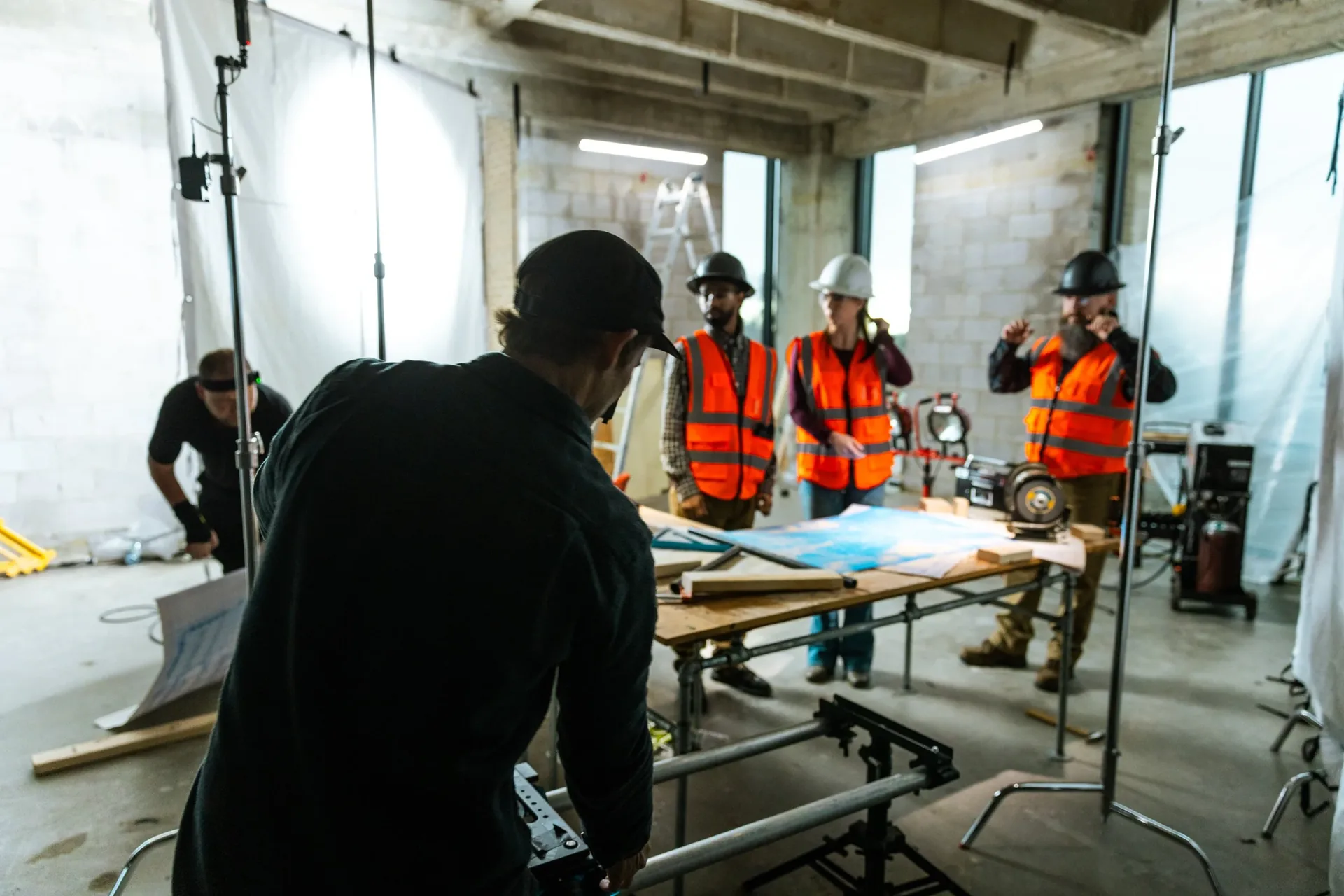 Wide behind-the-scenes view of the plans table: three actors in hard hats and vests wait at the table while the crew sets stands and diffusion