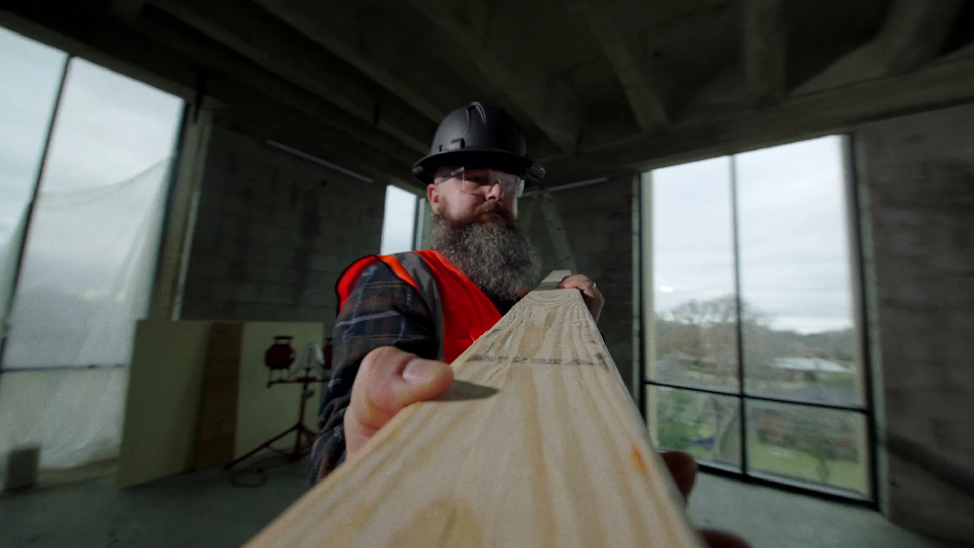 Delivered frame from a camera fixed to a 2x4: a bearded worker in a hard hat and safety vest carries the board through a room with tall windows