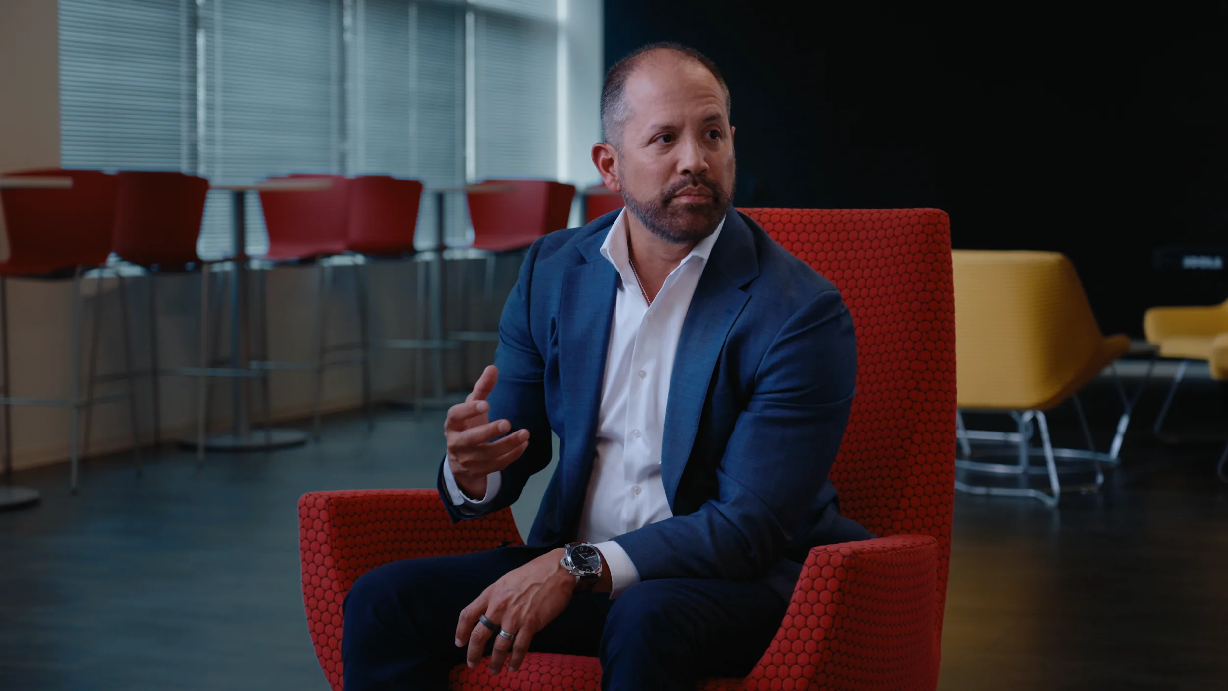 A man in a blue blazer gestures mid-answer from a red chair, bar stools and windows behind him