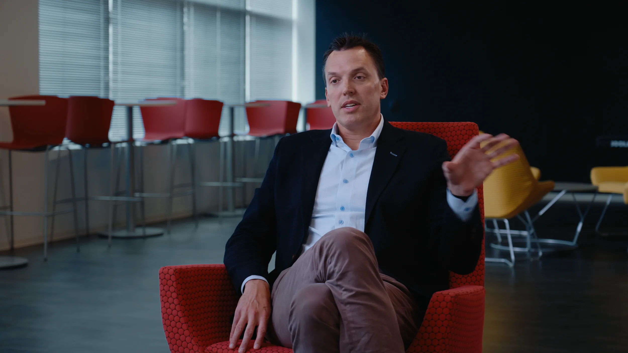 A man in a navy blazer speaks from a red chair, graded frame from the Houston interview series