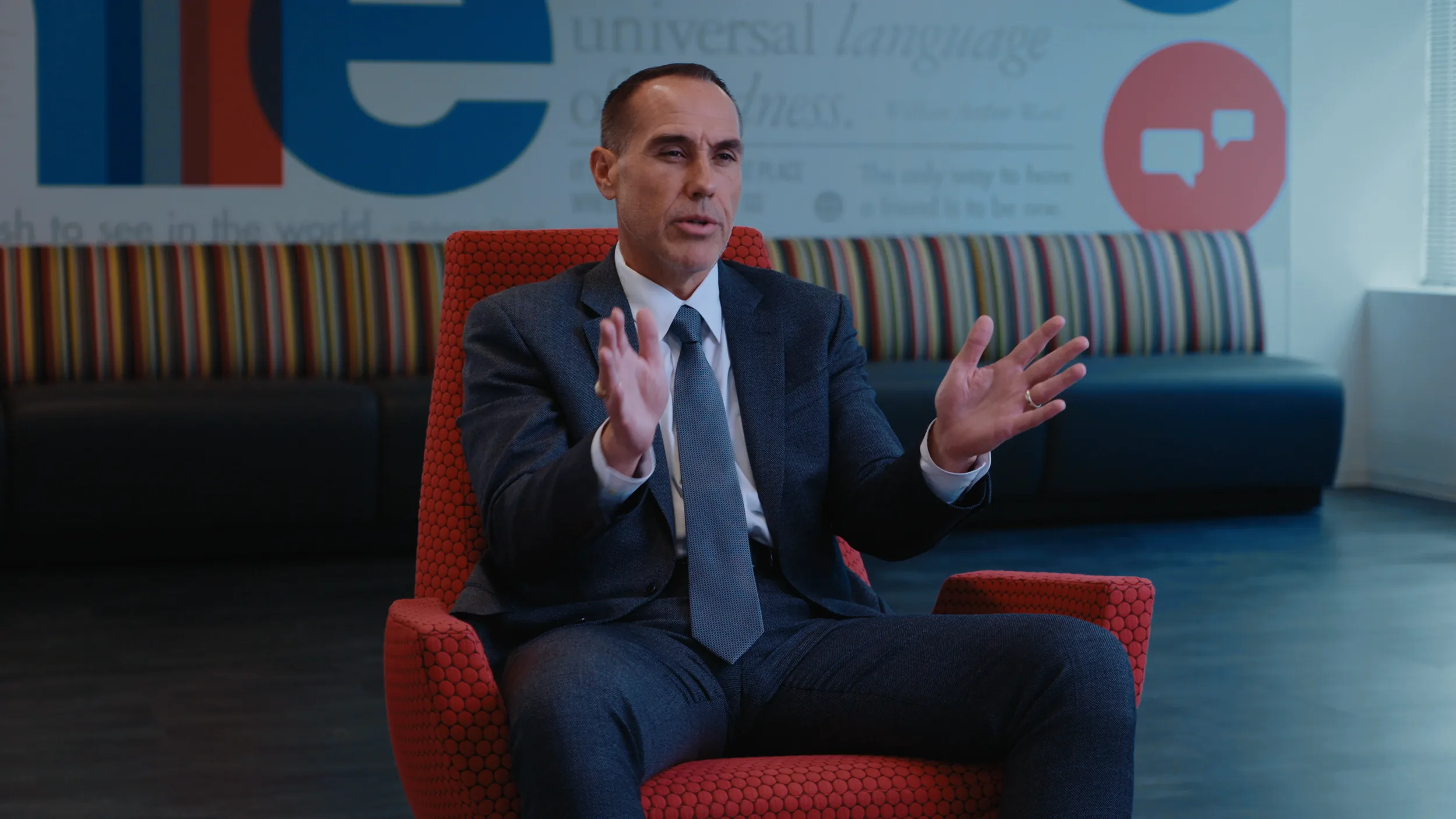 B camera: a man in a grey suit speaks from a red chair in front of a mural wall