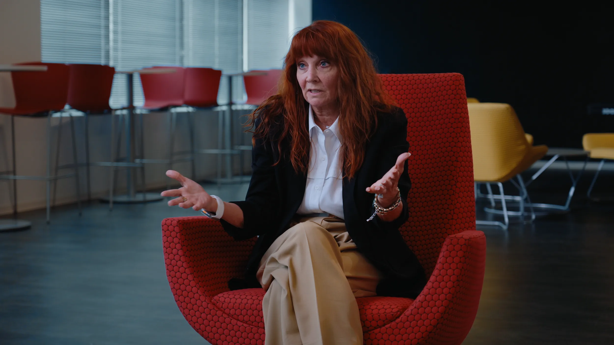 A woman in a black blazer speaks from a red chair, bar stools and windows behind her, Houston interview series