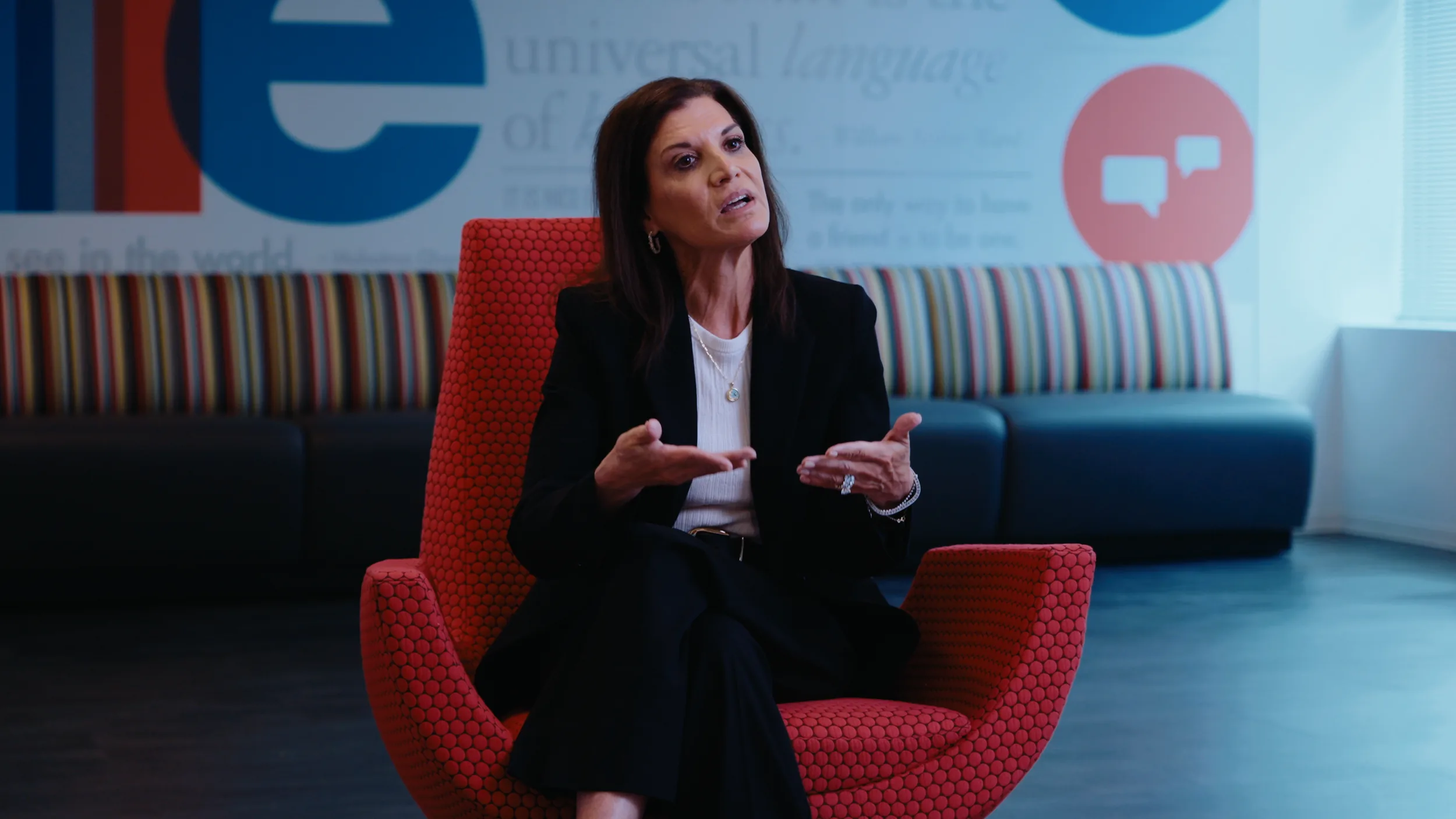 A woman in a black suit speaks with open hands in front of a mural wall and striped bench