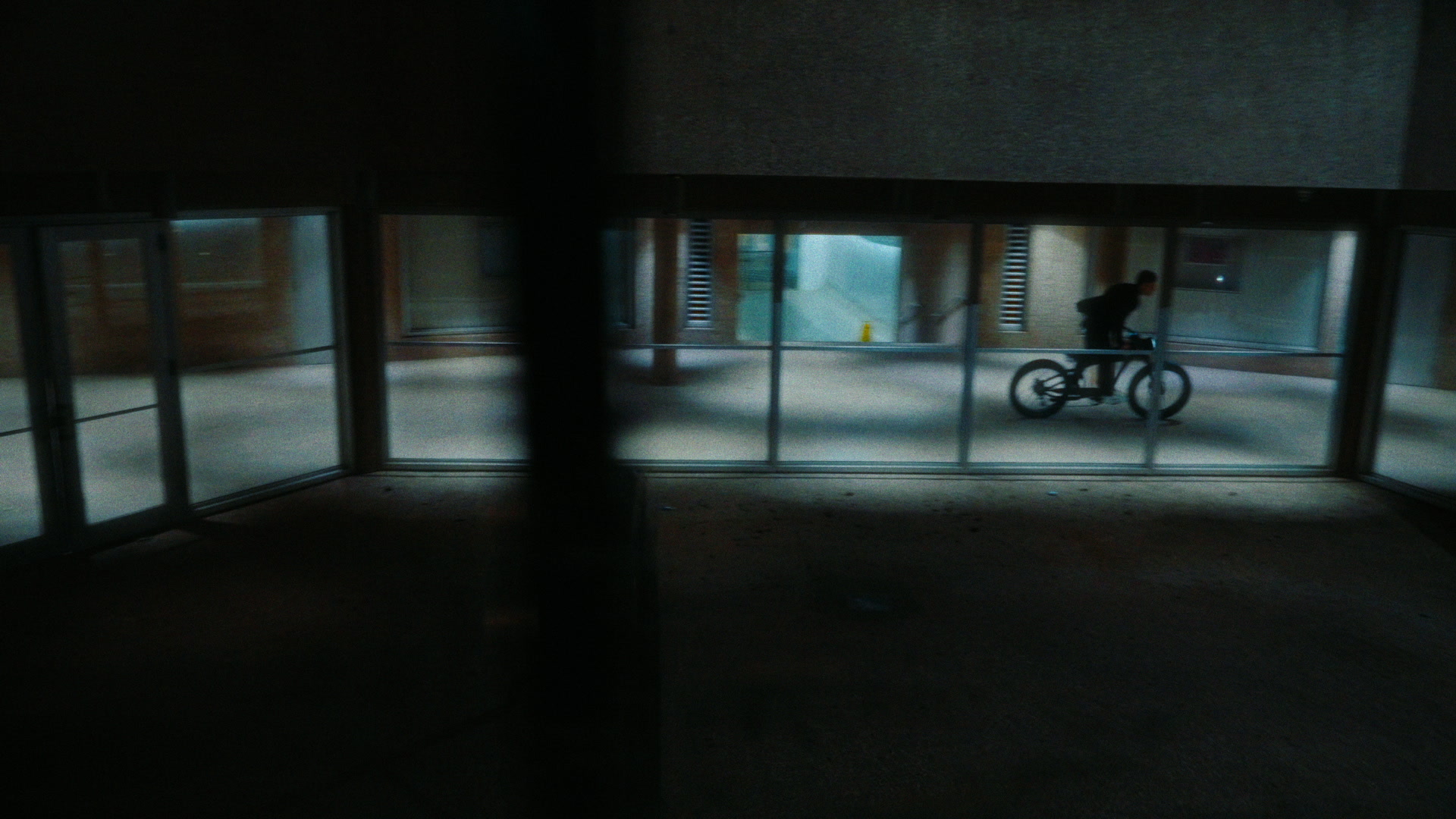 Night city plaza approach with bright bike headlight, blue practical spill, and exterior architecture.