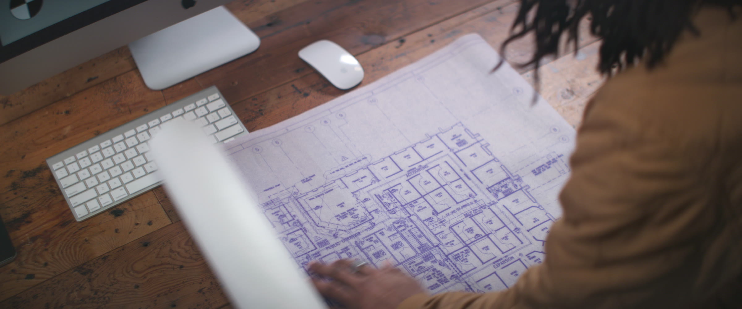 Overhead office planning frame with blueprint, keyboard, wood desk, and soft window spill.