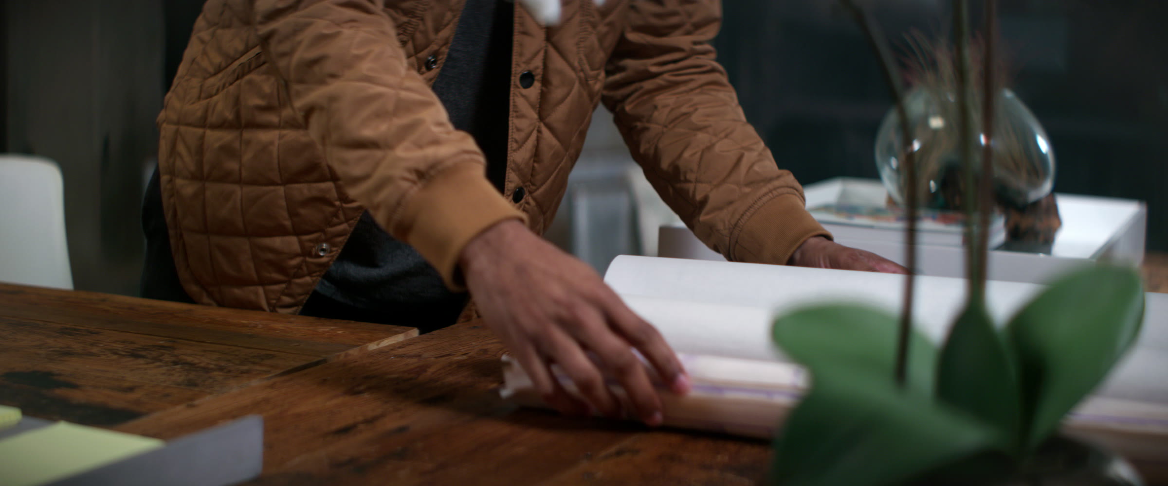 Hands moving rolled production plans across a wood table in a daylight office.
