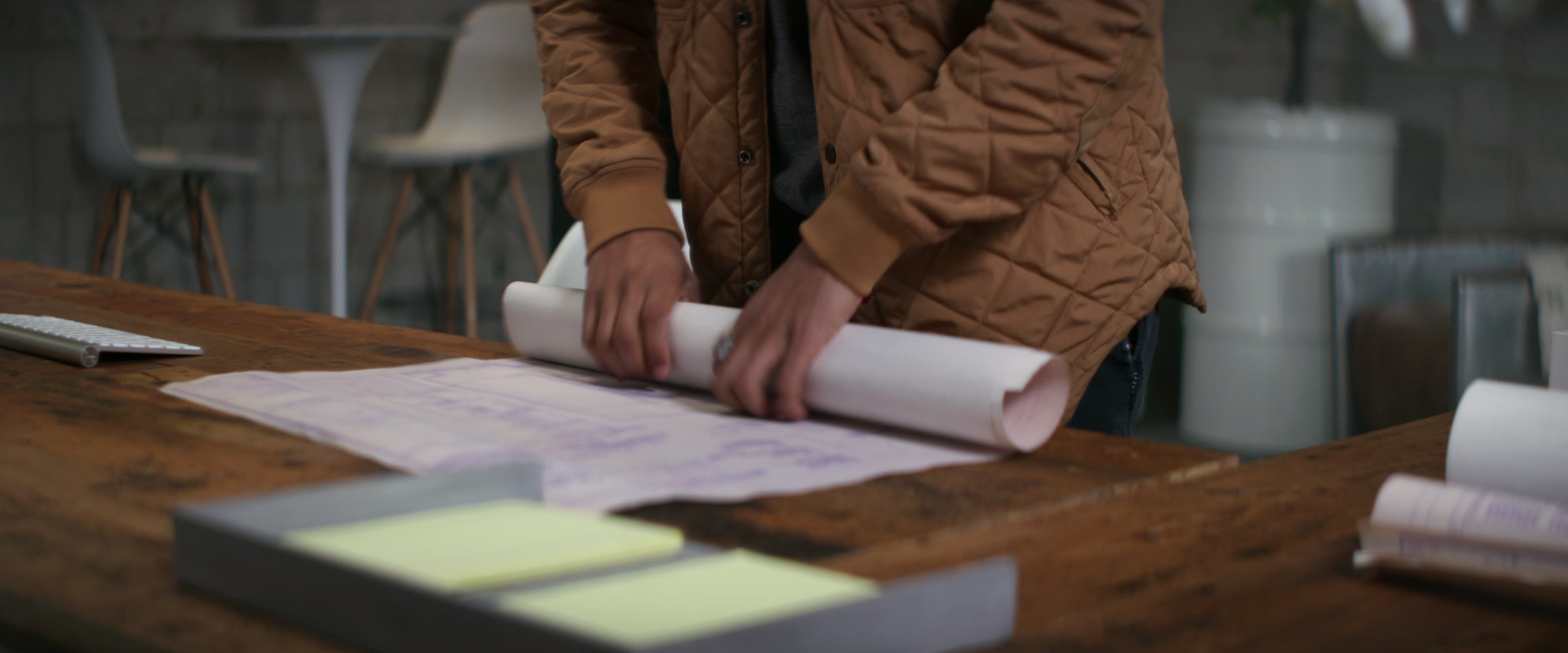 Close office tabletop frame with plans, paper roll, warm wood, and shallow focus.