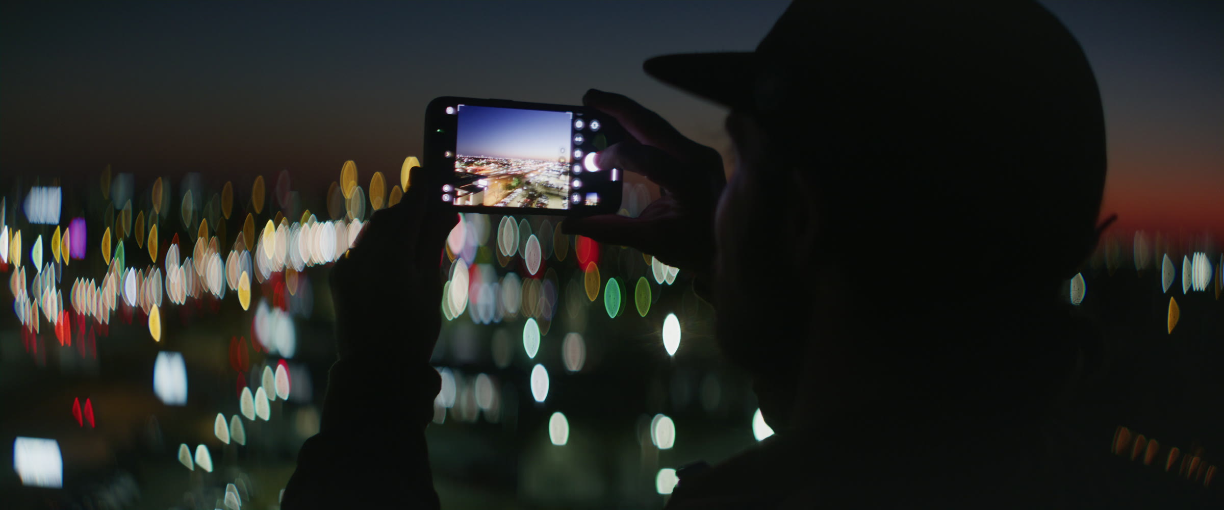 Phone filming city lights from a rooftop with colorful skyline bokeh in the background.