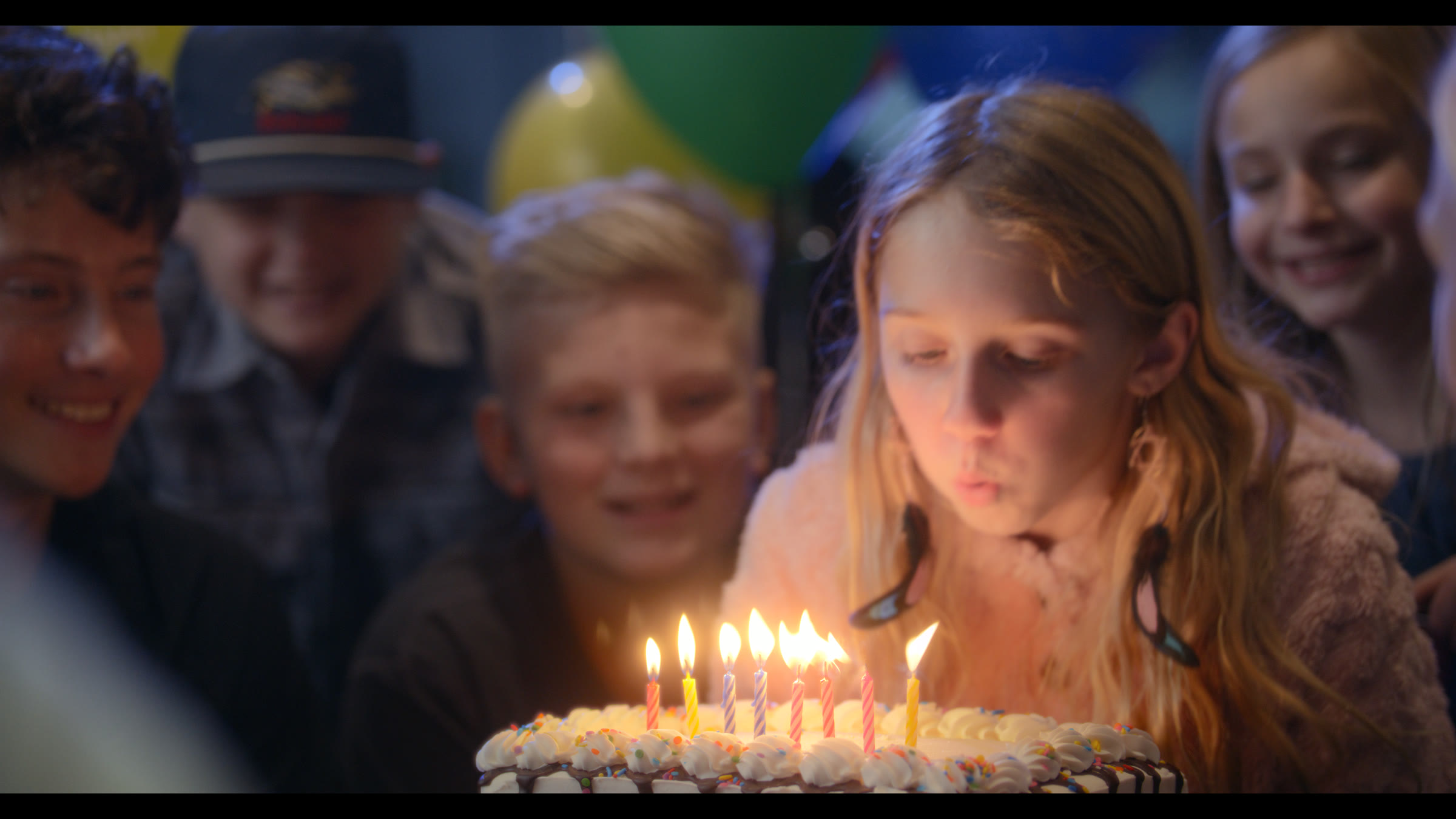 Birthday candle moment with a child centered in warm cake light and group reaction.