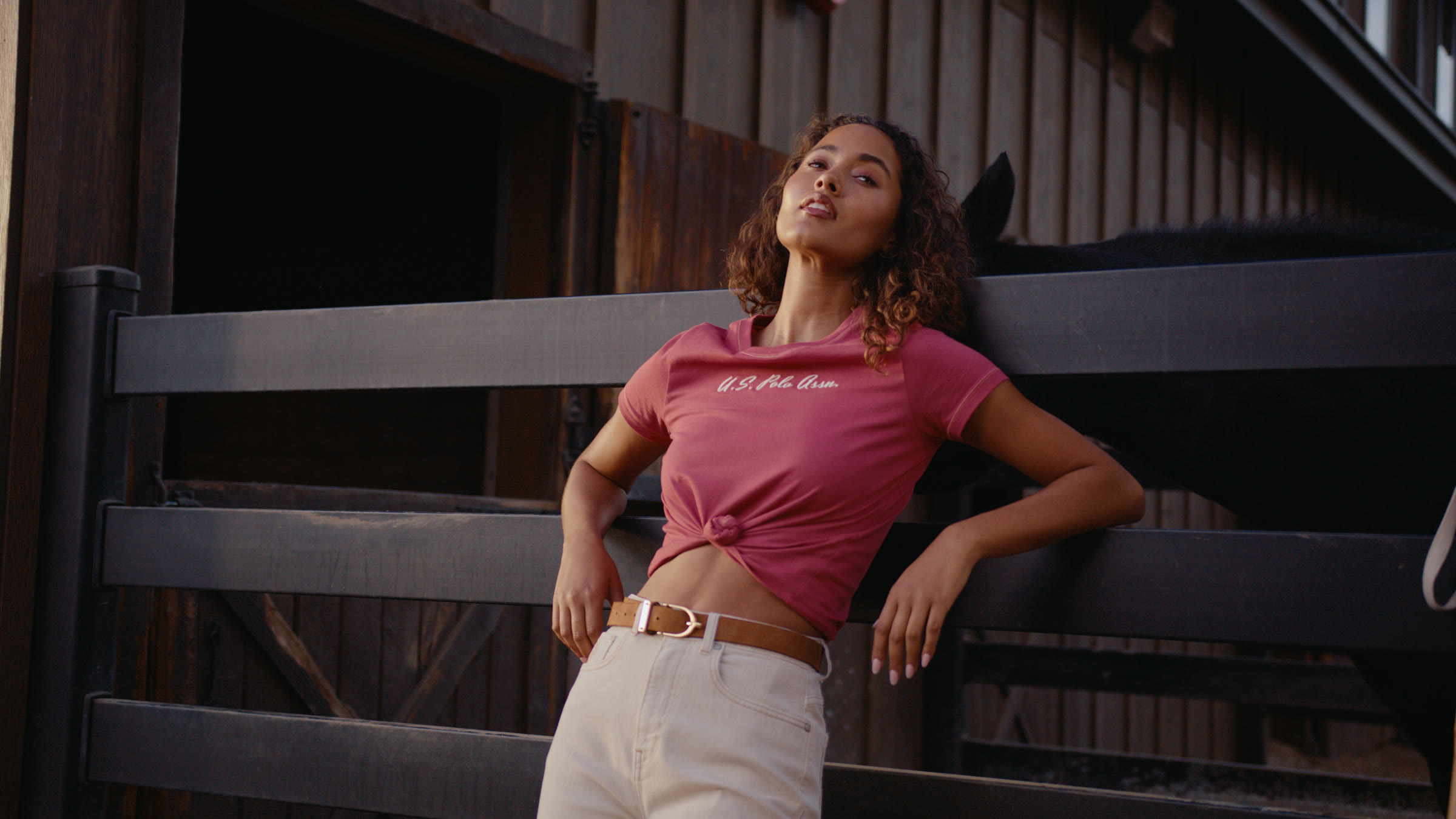 Model leaning against a barn rail in pink shirt with stable architecture behind her.