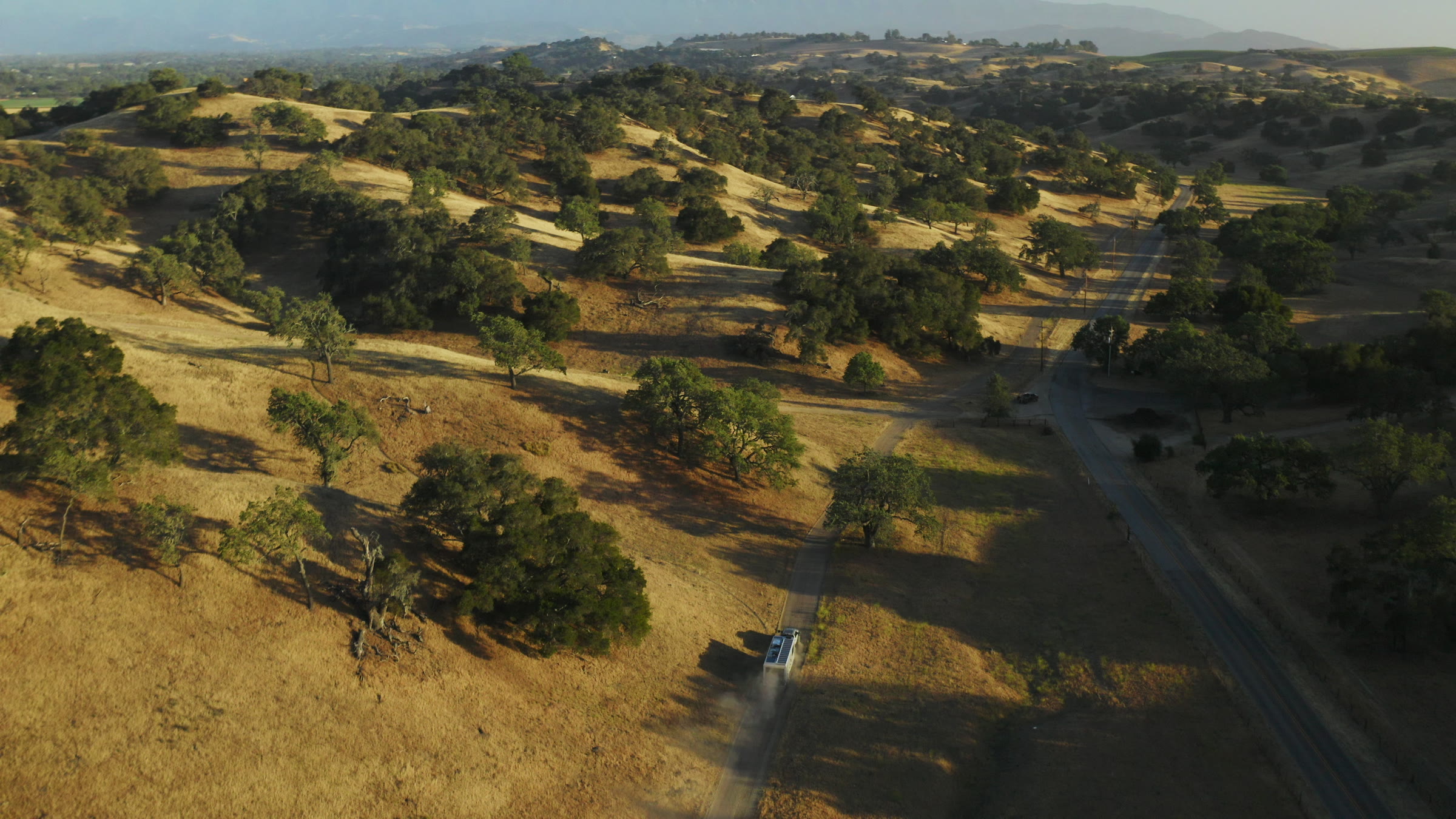 Overhead drone frame with oak-dotted hills, winding road, and warm late-day terrain.