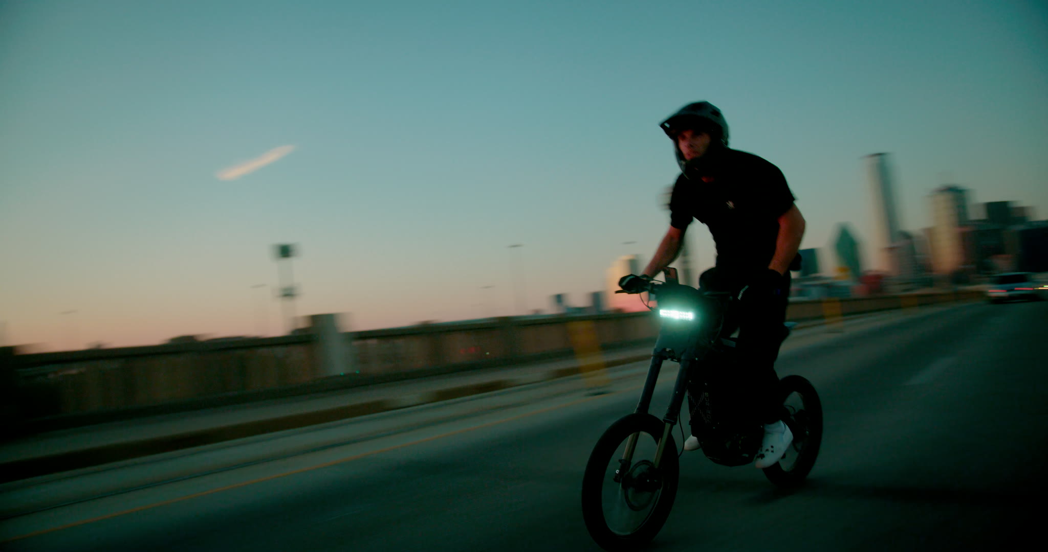 Bridge bike frame with headlight flare, teal road color, and downtown bokeh.