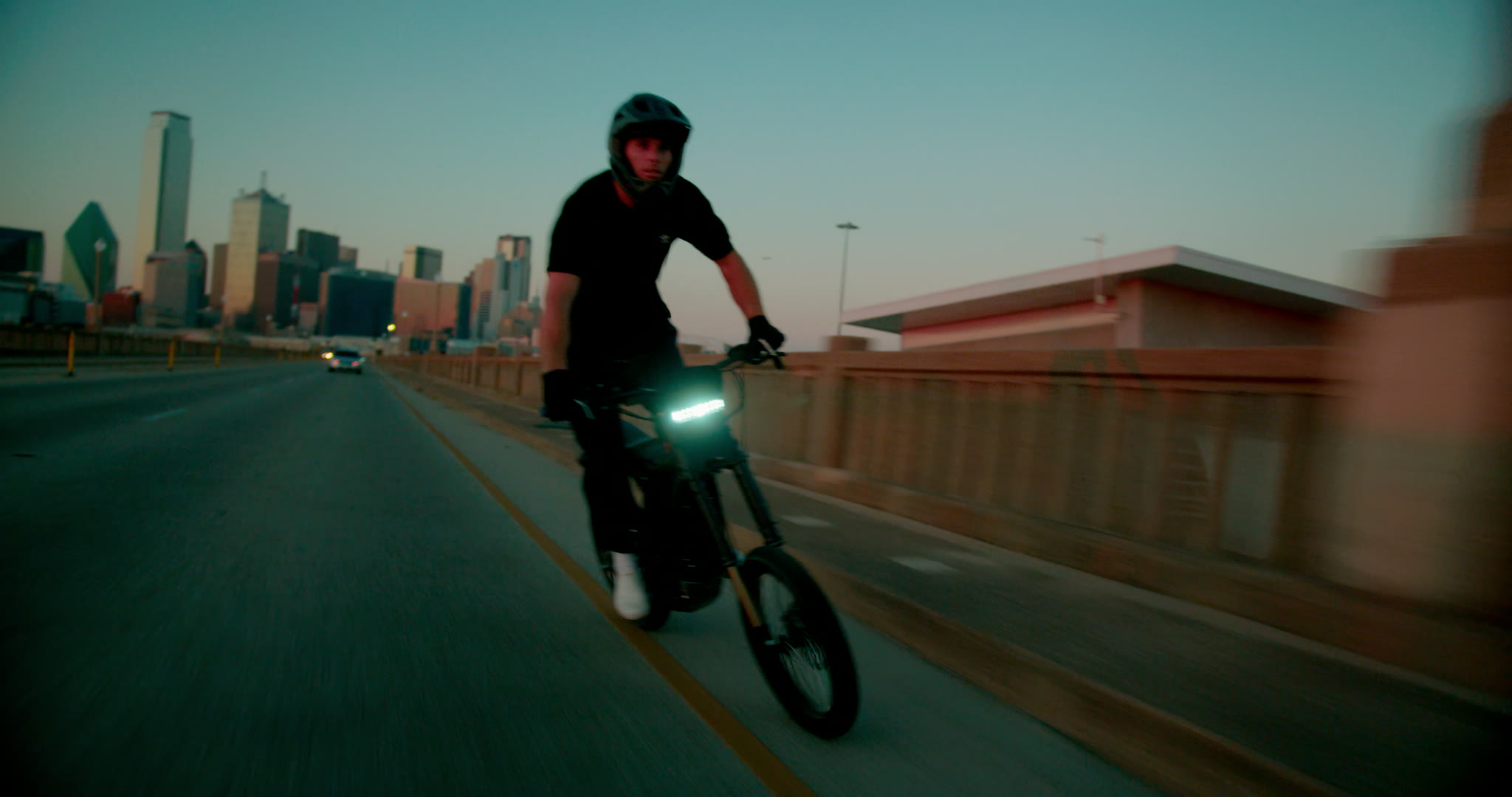 Wide bridge biking frame with Travis riding toward a downtown skyline at dusk.