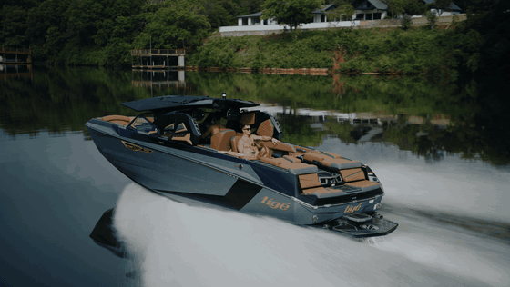 Looping Tige boat lakeside pass with passengers, a glassy wake, and wooded shoreline reflections.