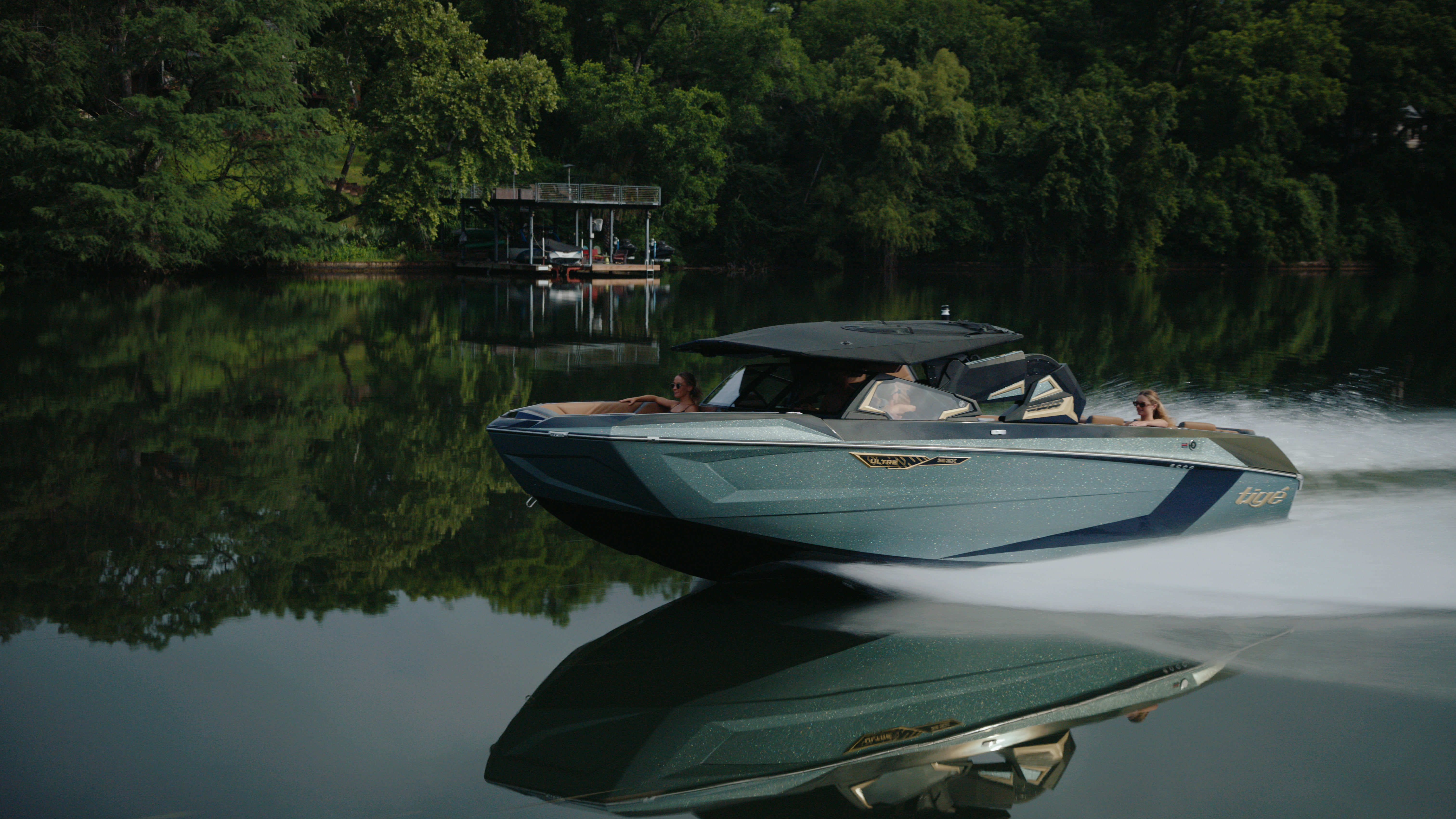 Side profile of a Tige boat moving across mirror-still lake water with a dark green shoreline reflection.